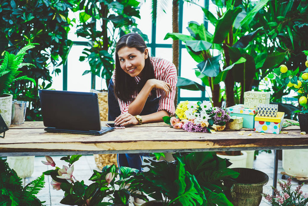 Portrait of asian female entrepreneur standing near desktop with laptop for checking mail from clients owning slower shop,prosperous proprietor of greenhouse satisfied with job looking at camera - Fotografie, Obrázek