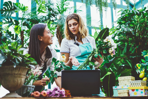  Young female florist discussing ideas for creating floral decoration due to online order checking on laptop,worker of flower retail store consulting with woman owner while making bouquet for sal - Foto, Bild