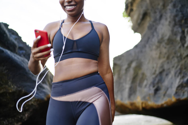 Cropped image of afro american female jogger dressed in stylish sport clothes for training listening music for workout in earphones connected to cellular.Sportive girl in active wear walking with phone - Photo, image