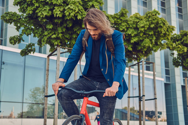 Handsome bearded male with long hair in stylish clothes and sunglasses with a backpack, riding a bicycle on the street, outdoors. - Photo, Image