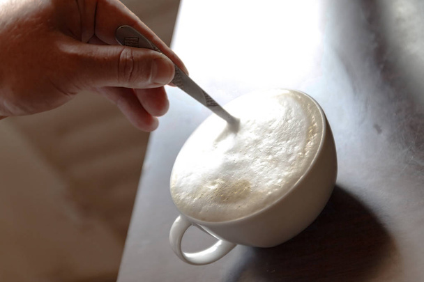 Cup of coffee with foam being stirred by a human hand - Foto, afbeelding
