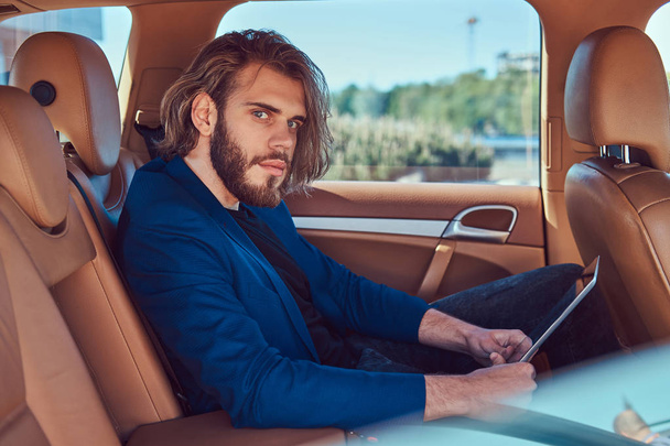 Handsome businessman with a beard and long hair sitting in the back seat of a luxury car and working with a tablet computer. - Photo, Image