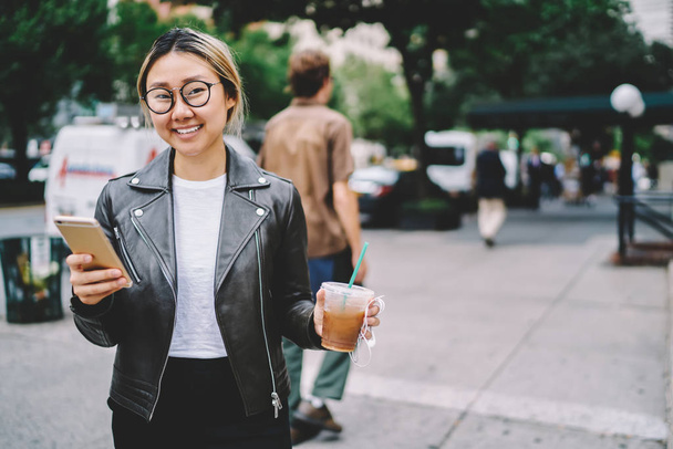 Smiling asian female tourist in eyewear walking on street in downtown using application on mobile phone for navigate, happy chinese girl sending message with coffee to go on street in city - Foto, Imagem