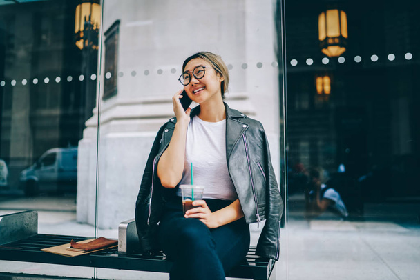 Cheerful Asian woman in eyewear having mobile phone conversation sitting on bus stop bench in urban setting, smiling Chinese hipster girl talking on cellular satisfied with cheap roaming tariffs - Foto, Imagem