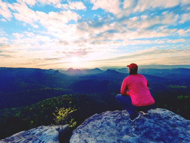 Turysta siedzący na skale. Człowiek w czerwony czarny ciepłe ubrania siedzi na klif i widokiem daleko.  - Zdjęcie, obraz