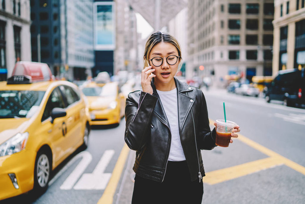 Young asian woman in eyewear walking with ice coffee cup calling taxi contact service operator on mobile,chinese trendy dressed girl having mobile conversation passing yellow cabs in downtown - Фото, изображение