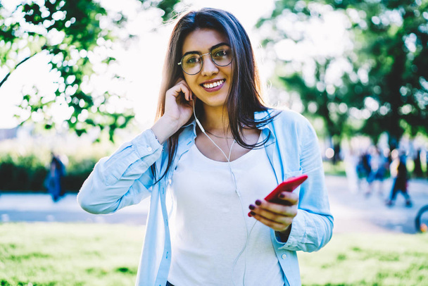 Half-length portrait of happy hipster girl in casual wear put on earphones for enjoy perfect song of favorite music,cheerful female student listening songs from playlist on smartphone looking at camera - Fotó, kép