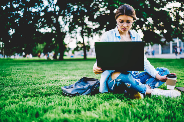 Thoughtful female freelancer working outdoors on laptop downloading files sitting copy space for advertising, pensive hipster girl in glasses doing remote job and installing application on netbook - 写真・画像