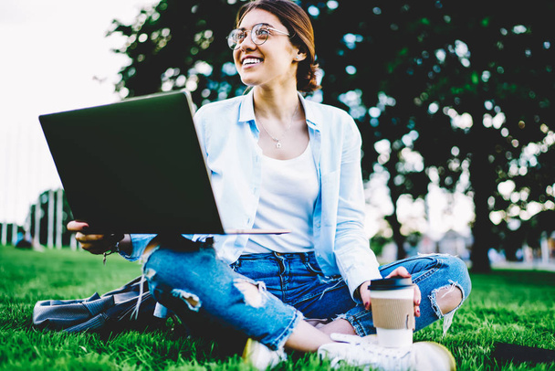 Happy young woman in eyewear having fun during coffee break sitting on green lawn in park, smiling female student satisfied with learning outdoors on campus using laptop for doing homework task - Photo, Image