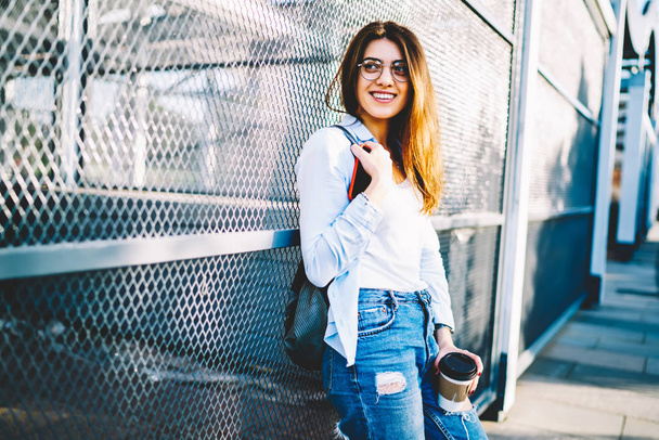 Cheerful young woman in casual outfit looking away standing on urban settings background, smiling hipster girl in stylish spectacles enjoying free time hold coffee to go having break outdoors - Foto, afbeelding
