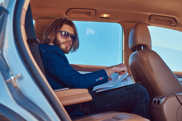 Красивый бизнесмен с бородой и длинными волосами сидит на заднем сидении роскошного автомобиля и работает с документами
. - Фото, изображение
