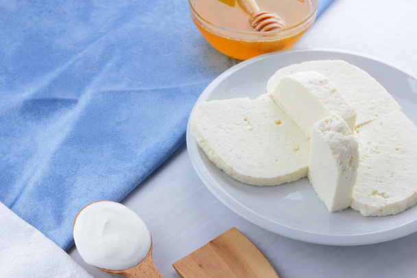 Τυρί, μέλι και ξινή κρέμα, φέτα σε λευκή πινακίδα, μαλακό τυρί σε λευκό φόντο, ξύλινο κουτάλι με ξινή κρέμα, γαλλικό πρωινό, μπλε πετσέτα, μέλι στα υαλικά, κενό για σχεδιαστής - Φωτογραφία, εικόνα