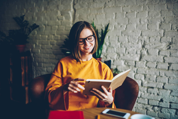 Pensive smiling young woman in eyewear reading interesting novel resting in coffee shop enjoying hobby.Charming student with book in hands spending free time on exam preparation in cozy cafe - Foto, Imagem