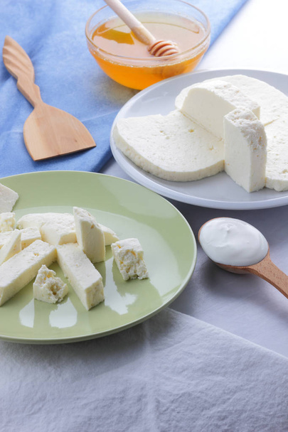 Μοτίβο τυρί, μέλι και ξινή κρέμα, φέτα σε λευκό και πράσινο πλάκα, μαλακό τυρί σε λευκό φόντο, ξύλινο κουτάλι με ξινή κρέμα, γαλλικό πρωινό, μπλε πετσέτα, μέλι σε γυάλινα σκεύη - Φωτογραφία, εικόνα