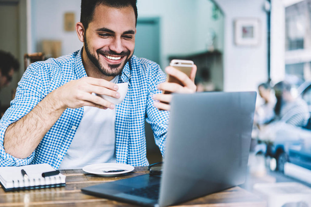 Cheerful freelancer enjoying coffee reading messages from friend in social networks working remotely in cafe,hipster guy happy about getting discount for online shopping checking mail on cellular - Foto, immagini