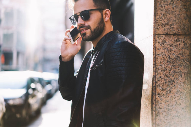Handsome male in trendy wear talking on mobile phone having cheap tariffs walking on city street,young hipster guy having telephone conversation with friend in roaming spending time outdoors - Photo, Image