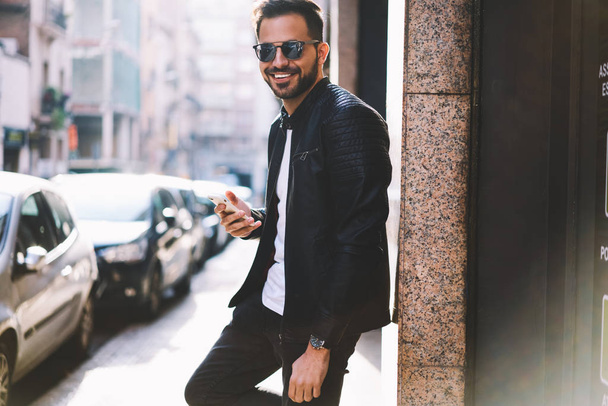 Half length portrait of cheerful hipster guy using smartphone standing on city street blogging in social networks,happy handsome male looking at camera chatting with friend online via telephone - Фото, изображение