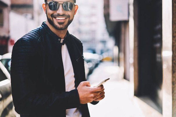 Cropped image of cheerful hipster guy looking at camera chatting with friend online via smartphone standing on city street,portrait of smiling male person sending multimedia messages via cellular - Valokuva, kuva
