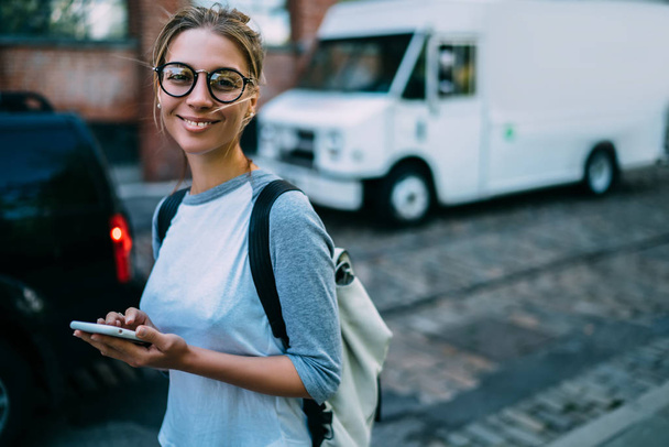 Portrait of smiling hipster girl holding smartphone chatting in social networks while walking on street,positive female traveler using application for transportation in city calling taxi onlin - Foto, imagen