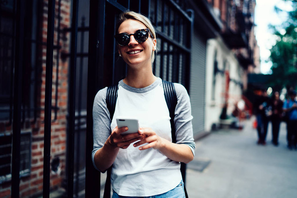 Happy hipster girl sending message via smartphone enjoying leisure strolling ,smiling female tourist in sunglasses satisfied with internet connection in roaming chatting online while sightseein - Фото, зображення