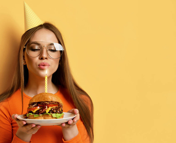 Woman with tasty big beef burger barbeque sandwich for birthday party with lit candle on pastel yellow - Photo, Image