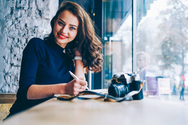 Porträt einer charmanten Studentin mit brünetten Haaren, die in die Kamera lächelt, während sie Notizen im Notizblock notiert, sitzend im Café mit Vintage-Kamera. Positive weibliche Aufnahmeinformationen in Notizbuch - Foto, Bild