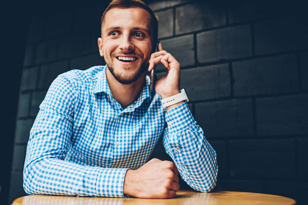 Cheerful hipster guy happy about getting good news during mobile phone conversation, successful businessman celebrating achievement of project excited while talking on telephone sitting in cafe - Fotografie, Obrázek