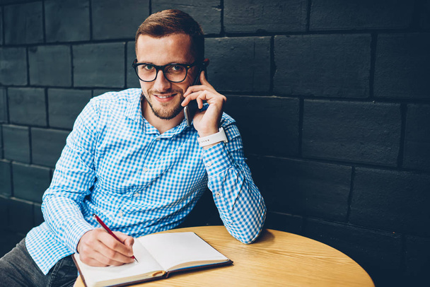 Portrait of smiling businessman noting information during phone conversation with colleague,positive male in spectacles talking with banking service operator writing in notepad looking at camera - Foto, imagen