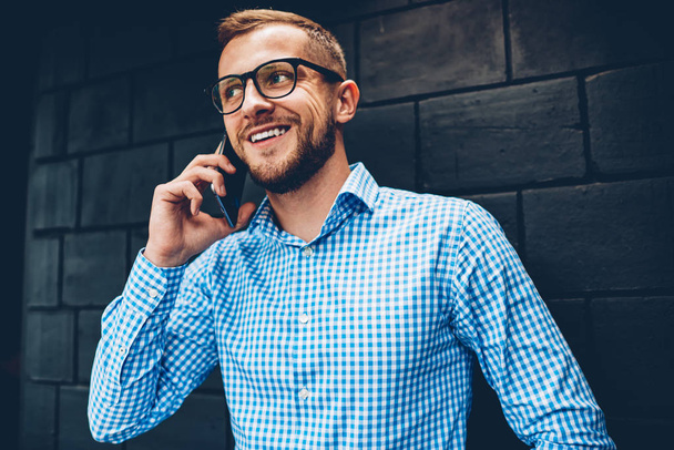 Smiling hipster guy in spectacles satisfied with tariffs for mobile phone calls spending time indoors,positive businessman exited with good news about project talking on telephone with colleague - Фото, зображення