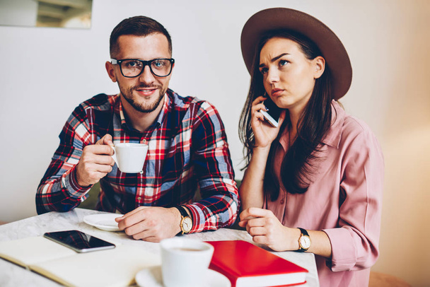 Young woman in trendy hat talking on phone while drinking coffee with her boyfriend in cafe, handsome man in eyewear looking at camera having meeting with female colleague making telephone call - Foto, immagini