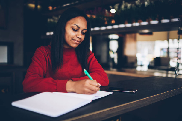Attractive young dark skinned model writing in notebook with blank pages spending time in cafe, Beautiful afro american businesswoman making financial notes planning budget for project in coffee shop - Foto, Imagem