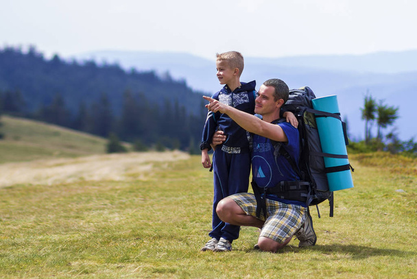 Father and son with backpacks hiking together in scenic summer green mountains. Dad and child standing enjoying landscape mountain view. Active lifestyle, family relations, weekend activity concept. - Photo, Image