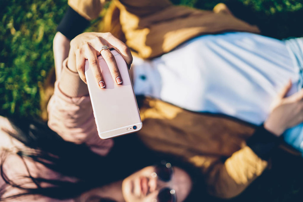 Top select focus on woman's hand holding modern digital telephone device and making selfie photos for social networks together with friend lying outdoors on green grass enjoying sunny weather - 写真・画像