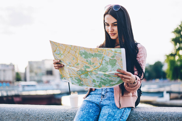 Positive young woman tourist with brunette hair holding map in hands and searching right destination sitting outdoors in urban setting on city promotional background with copy space area for text - Photo, image