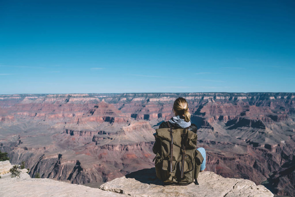 Back view of young woman with rucksack sitting on rocky cliff enjoying trip in USA exploring wild nature, female traveler fascinated with scenery of landscape with stone formation in National park - Фото, зображення