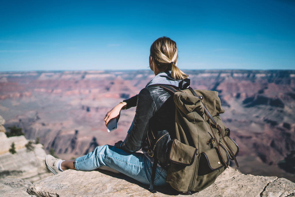 Young female tourist looking at beautiful scenery landscape of Grand Canyon sitting on peak hill,rear view hipster girl enjoying exploring wild environment of USA hiking in mountains with backpack - Zdjęcie, obraz