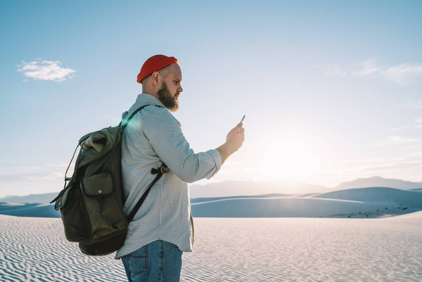 Serious male traveler with backpack exploring desert with white sand using navigation application on telephone, hipster guy wanderlust reading message on smartphone having good connection in roaming  - Photo, image