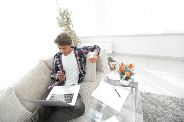 young man with laptop and smartphone sitting on sofa in living room. - Zdjęcie, obraz