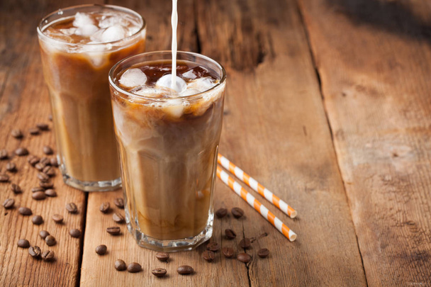 Ice coffee in a tall glass with cream poured over and coffee beans on a old rustic wooden table. Cold summer drink on a dark wooden background with copy space - Fotó, kép