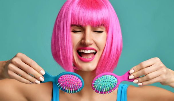 Closeup portrait of happy fashion brunette woman hold two small pink blue yellow small hair comb brush laughing in pink wig  - Fotografie, Obrázek