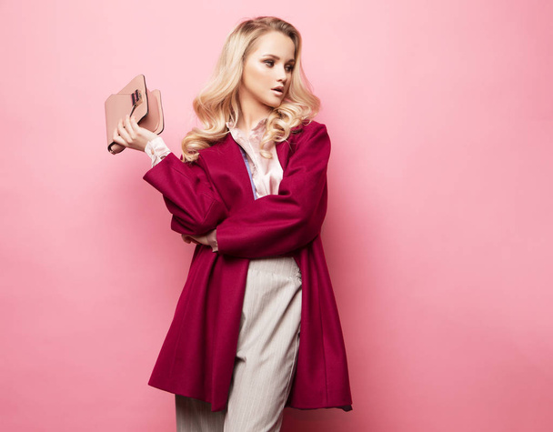 Beautiful blond woman in a blouse and pants wearing glasses, holding handbag posing over pink background. - Photo, image