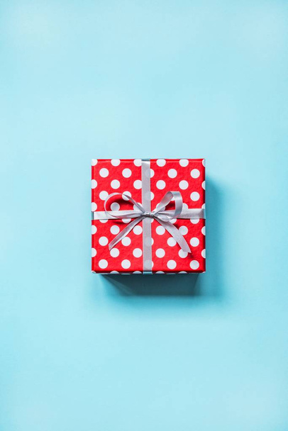 Κάτοψη του ένα κόκκινο διάστικτο δώρο κουτί δεμένο με ασημένια κορδέλα και φιόγκο σε μπλε φόντο.  - Φωτογραφία, εικόνα