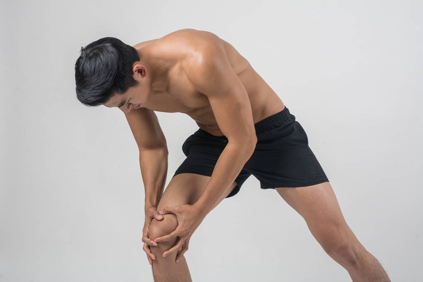 Νέος αθλητισμός άνθρωπος με ισχυρή γυμνασμένα πόδια κρατώντας τα γόνατα με τα χέρια του στον πόνο μετά από να υποστεί ζημία συνδέσμων απομονωθεί σε λευκό. - Φωτογραφία, εικόνα