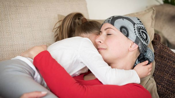 Young adult female cancer patient spending time with her daughter at home, relaxing on the couch. Cancer and family support concept. - Zdjęcie, obraz