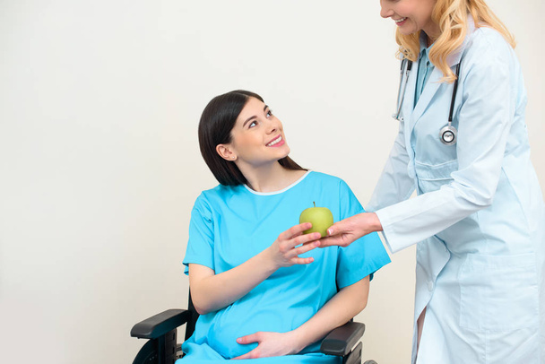 tiro cortado de ginecologista obstetra dando maçã verde à mulher grávida sorridente em cadeira de rodas
 - Foto, Imagem