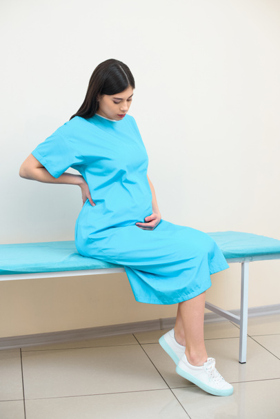Mladá těhotná žena sedící na posteli v nemocnici a drží ji zpět - Fotografie, Obrázek