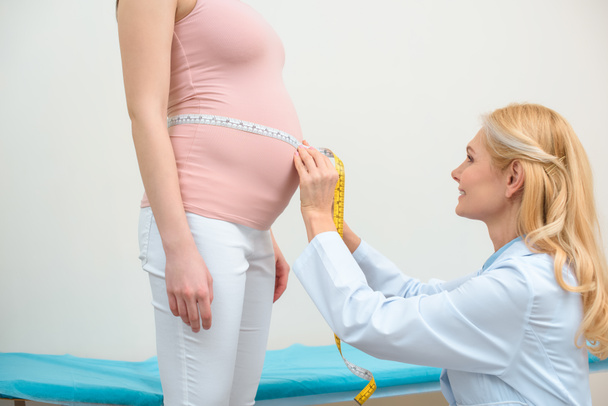 вид сбоку акушера-гинеколога, измеряющего размер живота беременной женщины с помощью измерительной ленты
 - Фото, изображение