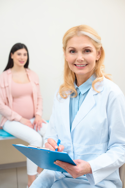 акушер-гинеколог с планшетом и молодая беременная женщина размыта на заднем плане
 - Фото, изображение