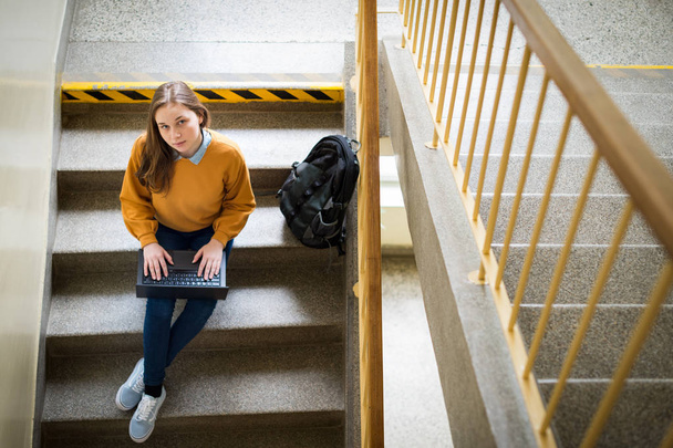 Young female college student sitting on stairs at school, writing essay on her laptop and looking up at the camera. Education concept. View from above. - Фото, изображение