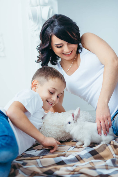 Family portrait of the happy smiling mother and her cute little blonde son playing with lovely fluffy rabbit while lying on the plaid at home. - Fotoğraf, Görsel